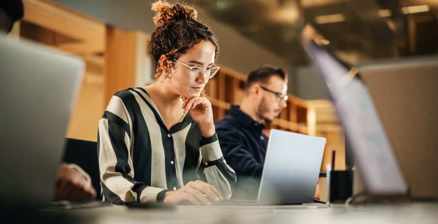 Portrait of Enthusiastic Hispanic Young Woman Working on Computer in a Modern Bright Office. Confident Human Resources Agent Smiling Happily While Collaborating Online with Colleagues.